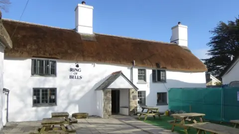 David Smith/Geograph The picture shows a traditional English pub with a thatched roof and whitewashed stone walls. The building has small grey-framed windows. The pub’s name “Ring of Bells” is visible on the wall near the entrance, which has a stone porch. In front of the pub, there is a courtyard with wooden picnic tables and benches.