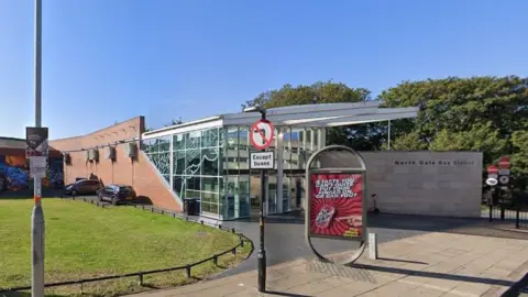 Google Glass-fronted building with stone wall adjacent bearing the legend "North Gate Bus Station". The rear of the building is faced in brick. There is a free standing advertising display stand on the pavement in the foreground promoting a soft drink, and a sign indicating no left turn except for buses. A grass plot is visible to the left.