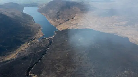 NI Water A burnt mass of land is in the foreground. It is clearly black with smoke rising from it. In the middle of the picture is Silent Valley reservoir.