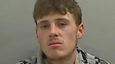 A blurry mugshot of Bradley Thomas who has short, brown hair and stubble on his upper lip and chin. His lips are pursed and he is wearing a grey and black patterned top.