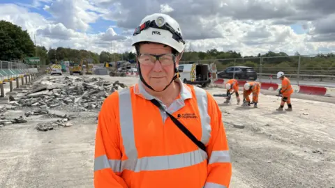 A head and shoulders shot of Terry Robinson. He is wearing a fluorescent orange shirt with a collar. He is also wearing safety glasses and a hard hat with a torch on the front. Behind him is the over bridge on the A40. Three workers are immediately to his right in the background hitting and drilling the concrete on the bridge. There's a white van with its double doors open over Robinson's right shoulder with a car passing through the single lane of traffic set up on the other side. To his left is a large pile of broken up concrete rubble. In the background there are various other construction vehicles, a green tree line behind that and a cloudy sky streaked with some blue spots.