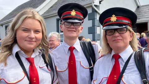 Three people dressed in white and red band uniforms pose for a photo at the Rossnowlagh parade. A young man is standing in the middle wearing a black peaked uniform cap with a red an gold trim, black square-framed glasses, a white shirt and a red tie. Two women with blonde shoulder-length hair are standing on either side of him smiling. They are similarly dressed but the young woman on the left is not wearing a cap. 