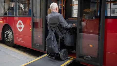 A person in a motorised wheelchair wearing grey suit boards a bus in London using a wheelchair ramp in October 2022