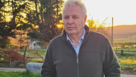 John Kirk John Kirk, a man with white hair, is wearing a blue shirt and brown fleece jacket. He is standing in a garden with golden sun behind him.