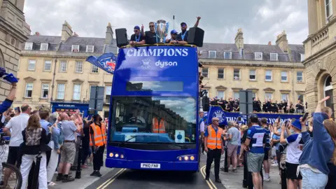 Blue open top bus with Bath Rugby players on top holding a large silver trophy drives through the city. 