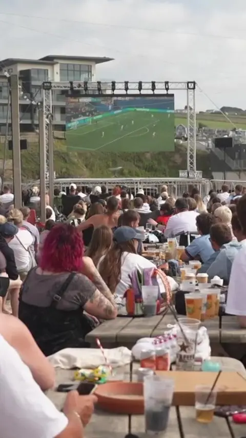 A crowd of people sitting at tables on a beach watching a big screen showing football.