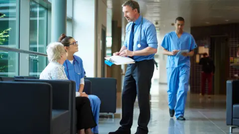 sturti A hospital setting. There are five people in total, four in the foreground and one in the background. Two of the people in the foreground are sat on chairs. One is in hospital scrubs, while the other is not. They are both women. One has short white hair, while the other has long brown hair and glasses. They are both looking up at a man wearing a blue shirt and tie. He is holding a clipboard and has a stethoscope around his neck. The fourth person in the foreground is a man in hospital scrubs with dark hair. He is walking past. The person in the background has their back to the camera.