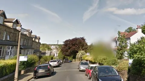 Google A residential street with parked cars on both sides of the road. Many of the front gardens are lined with shrubs and trees.