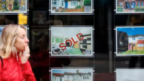 An estate agent's window showing a house being advertised says 'SOLD' in red across it. 