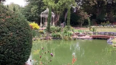 Avenues Open Gardens A large pond is surrounded by planting and a white obelisque structure. A little bridge crosses the stream and purple thistles are in the foreground.