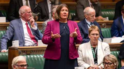 A woman wearing a purple jacket and navy blue dress with short brown hair stands in a room filled with other people wearing smart clothing. 