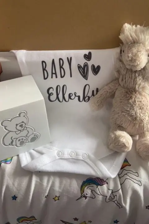 Lucy Ness A cubed box, with a teddy bear picture on the site, sits next to a white babygrow with "Baby Ellerby" written on it. Also pictured is a soft toy.