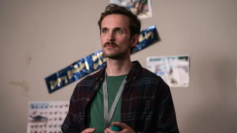 BBC/ Conker Pictures Vittorio Angelone in Rewarding. He has brown hair and a moustache and beard. He is wearing a green t-shirt, a burgundy checked shirt over the top of it and a grey lanyard. 