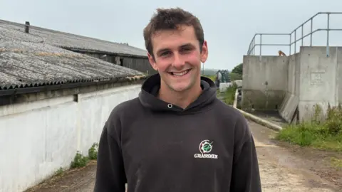 A person in a dark hoodie stands smiling on a farmyard lane between corrugated barns and a large concrete agricultural structure, with grass and equipment around under a grey sky.