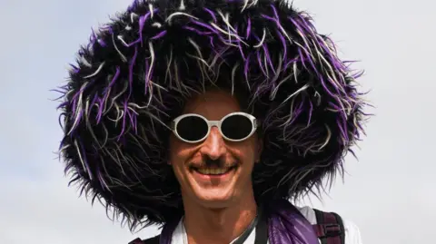 Reuters A man smiles at the camera. He wears round, white-rimmed sunglasses and a huge black, purple and white furry hat.