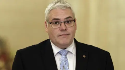 PA Media Gavin Robinson standing in front of a blurry background. He is wearing a black suit and blue tie, and is wearing glasses.