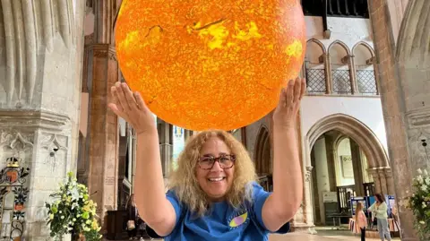 A woman with shoulder-length blonde hair is smiling and has her arms up in the air, creating the illusion that she is holding the large model of the Sun which is above and behind her. She is wearing tortoiseshell glasses and a blue top with an eye logo on it. She is standing inside a church.