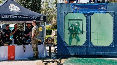 Royal Navy To the right is a large navy blue dive tank with two large windows. The tank is filled with water and one diver wearing diving equipment is seen through the left window. To the left is a Royal Navy stall with dive equipment on display.