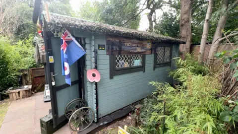 A side view of a green garden shed, which has a red poppy and RAF flag pinned to the outside and a banner saying Victory in Europe draped along it. It stands on flagstones in a garden surrounded by trees.