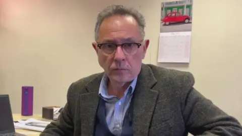 Geoff Ryall-Harvey looking straight at the camera. He is a man in his 60s wearing a green-grey tweed jacket, blue waistcoat and open-necked blue and white striped shirt. He's wearing glasses with frames which are darker at the top than the bottom and is in an office with magnolia walls and a calendar on the wall and a laptop on the desk behind him.