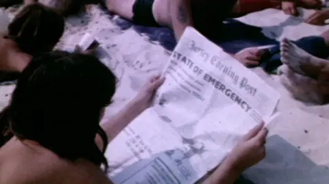 People dressed in swimsuits are lying on the beach reading. a woman is looking at the front page of the Jersey Evening Post with the headline States of Emergency.