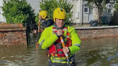 Two men wearing yellow helmets and flood gear wading waist-deep in flood water