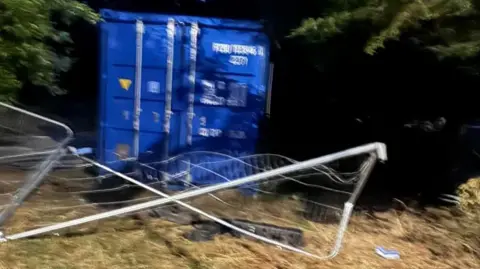 Blue shipping container with crushed metal fencing on the ground in front.