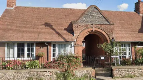 A street view image of a small house made of red bricks. There are plants and bushes outside the home with steps leading to a porch and entrance.