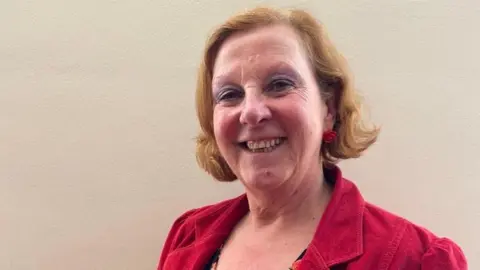 Lyn Sharpe A woman with mid-length ginger hair and wearing a red coat, smiles while standing in front of a wall inside.