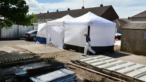 PA Media White tents in a residential car parking area being searched by forensic officers. The car park has a gravel surface with yellow and black tape across part of it. It lies in front of a row of houses