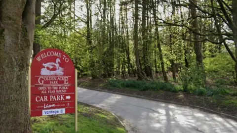 A wooded area with a footpath running through. There is a red wooden sign which reads 'Welcome to Golden Acre Park Car Park'.
