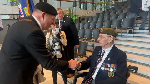In a conference hall with tiered seats a man in dark military uniform and beret leans down to shake the hand of an elderly man with glasses who is also in uniform with a beret and wearing medals. A young woman with glasses looks on and films on her mobile phone.