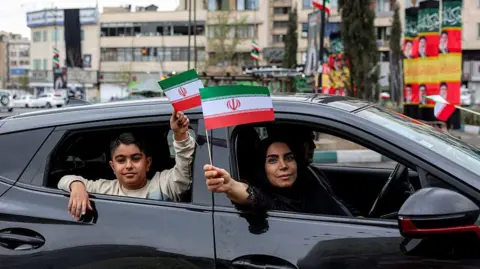 AFP via Getty Images A dark-haired woman and a young boy in a sweatshirt wave small Iranian flags from the windows of a black car. The flag is a horizontal tricolour with green, white and red stripes, and an Islamic central emblem. Photo: 25 March 2026
