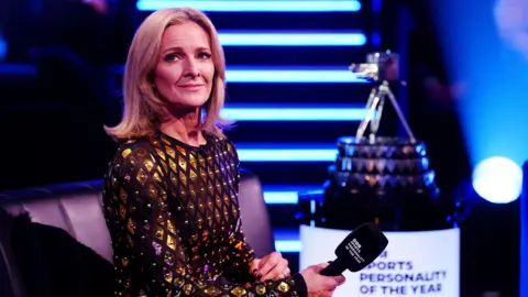 PA Media Gabby Logan wearing a black dress with gold sequence scattered across it. She is holding a microphone in her right hand and the Sports Personality of the Year award can be seen blurred in the background on her right.