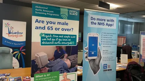 Two signs show advice on how to use the NHS app and advertise a scan for men over aged 65.