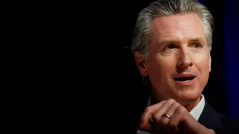 Gavin Newsom speaking at an event wearing a white button-down shirt and suit jacket