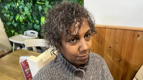 John Devine/BBC Navlett Cooper, has dark frizzy hair and a greyish woollen jumper done up above her neck, she is sitting at a table in a café with pine wooden walls and a menu on a table visible behind her.