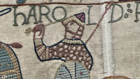 A close up of the Bayeux tapestry shows a soldier holding an arrow in one hand, and a blue shield in the other