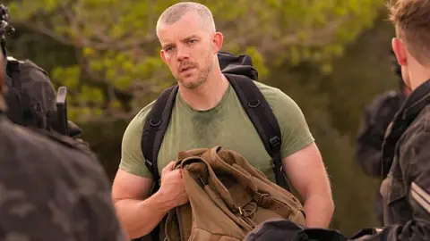 BBC Studios/Bad Wolf/Alistair Heap Russell Tovey is in character as Barclay in The War Between. He is outside, stood in front of a backdrop of green trees. He is broad, wearing a green t-shirt with black rucksack straps over his shoulders. He is holding a brown bag and surrounded by what looks like a group of military staff in uniform.