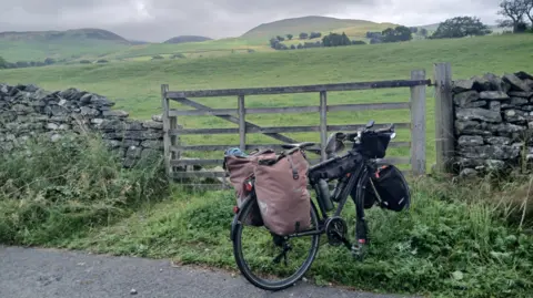 Chistoph Held A black bike with bags attached to the rear wheel is standing next to a wooden fence set in a stone wall. Beyond the fence are rolling green hills with bleak mountains in the distance under an overcast sky.