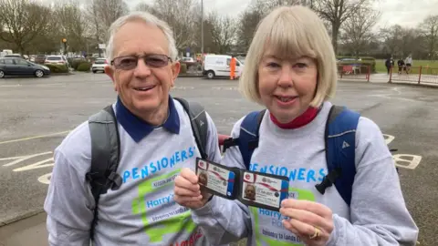 Martin Heath/BBC Nick and Kerstin wearing grey sweatshirts labelled "Pensioners' Pilgrimage" and "Harry's Pals". Nick has short white hair while Kerstin has short blonde hair. They are both wearing backpacks. Kerstin is showing their bus passes to the camera. There is a large bus turnaround area in the background.