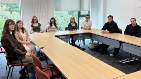 Eight classmates sat around a rectangular formation of chairs and tables 