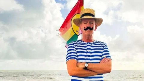 Normal Cook, AKA Fatboy Slim, dressed as a Victorian-era swimmer in a blue and white button up swimsuit with short sleeves. He is wearing a straw hat with a black band around it and a fake handlebar moustache. Behind him is a rainbow flag with a yellow smiley face on it blowing in the breeze.