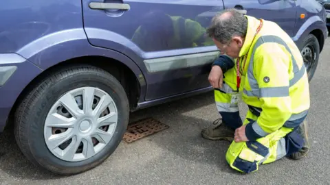 Northamptonshire Fire and Rescue Service A man kneeling down, wearing a yellow high-vis outfit, and inspecting a hydrant which has been parked over by a blue car.
