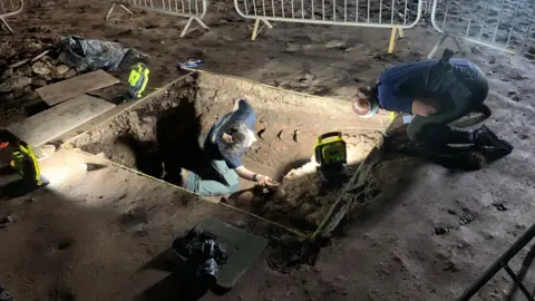 Archaeologists at a site are digging in a rectangular hole in the ground surrounded by metal fences. They are wearing head torches