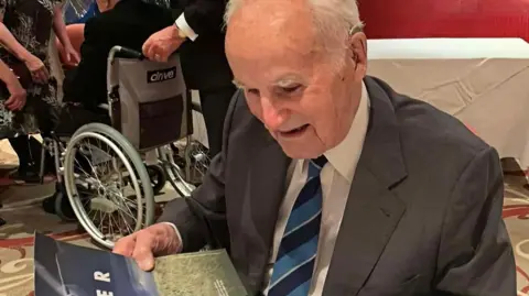 Jack Dark wears a dark jacket, white shirt and blue striped tie, as he looks at a photograph at a social event.