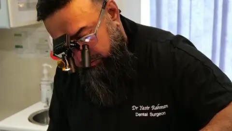 Instagram A middle aged Asian man, with a long black beard streaked with grey and black hair, treats a patient who is out of frame. He has a black dental uniform with 'Dr Yasir Rahman Dental Surgeon' stitched on to the chest in white lettering. 