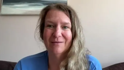A woman called Emma Hookway. She is sat on a sofa in front of a grey wall with an artwork on the wall. She has long fair hair and is wearing a blue t-shirt.
