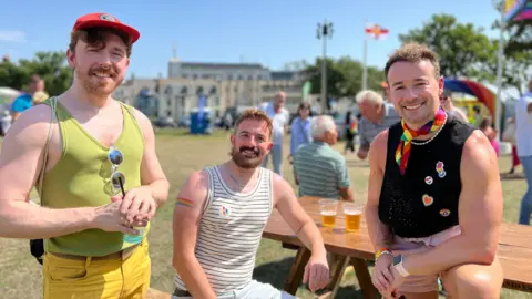 Sam, Alex and Liam are gathered around a wooden picnic table outdoors on a sunny day. Sam wears a red cap, a green vest and yellow trousers, Alex wears a striped tank top and shorts, and the Liam wears a black sleeveless shirt with enamel pins and a rainbow scarf. Plastic cups of beer are on the table. In the background, people are sitting or walking, with flags and trees visible under a clear blue sky.