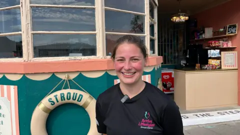A woman standing outside a poolside kiosk wearing a black Active Lifestyles Stroud District T-shirt. Behind her is a cream lifebuoy marked “Stroud 1937”, with the kiosk entrance and snack displays visible in the background.